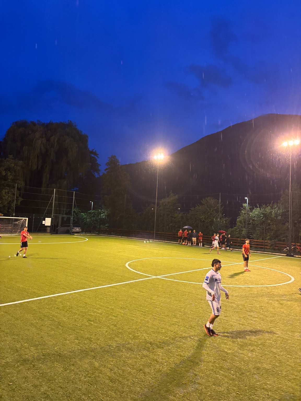 Fußballspiel unter Flutlicht im Regen vor einer Bergkulisse