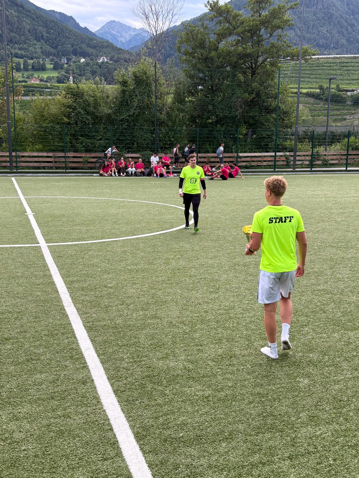 Turnierszene auf dem Fußballplatz: ein Pokal in den Händen eines Staff-Mitglieds, dahinter Spieler und Berglandschaft